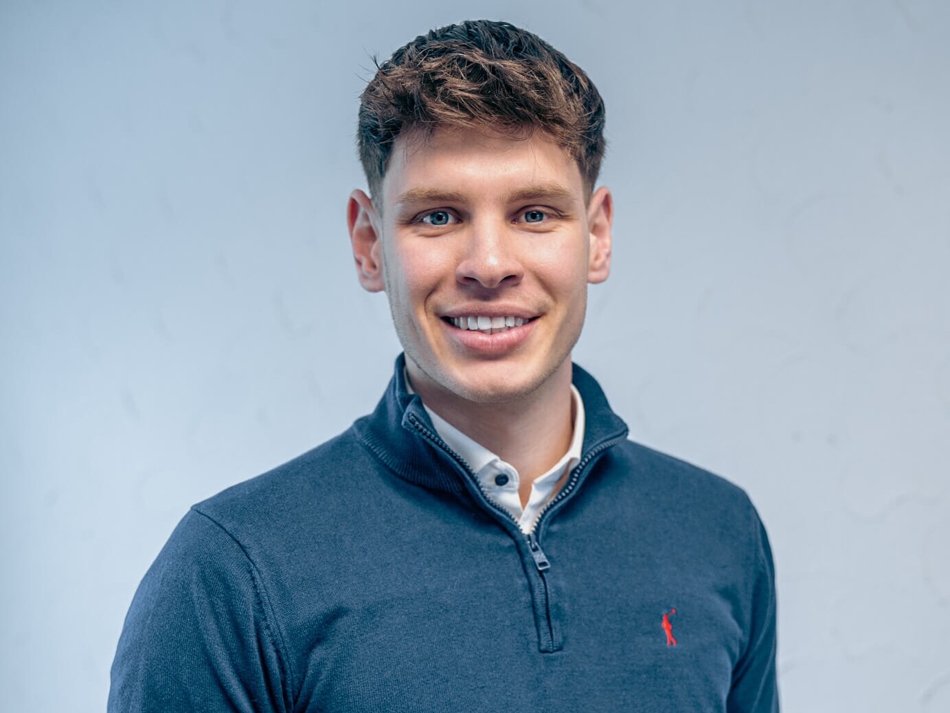 Junger Mann mit dunkelblauem Pullover l&auml;chelt vor neutralem Hintergrund