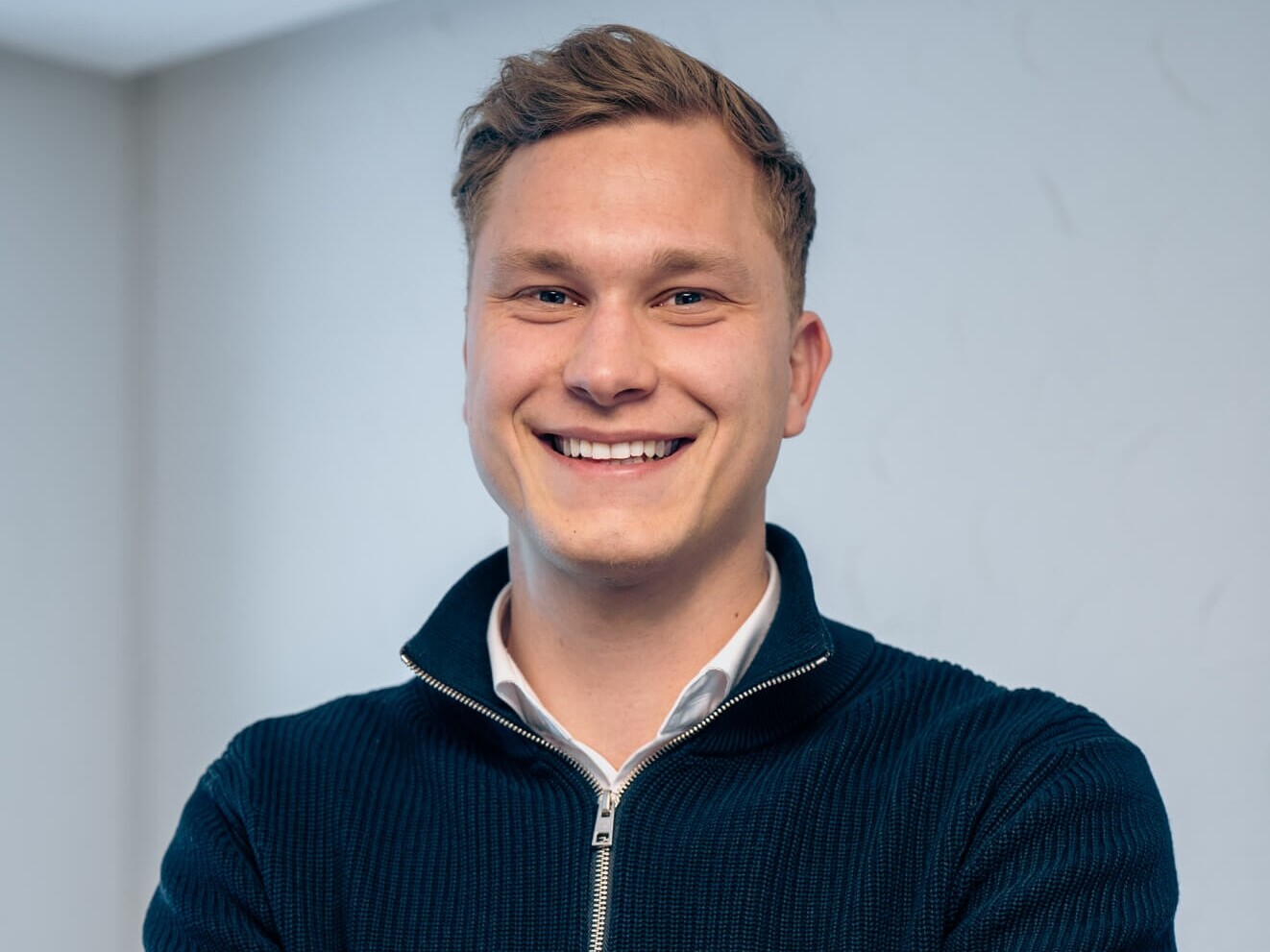 Junger Mann mit dunkelblauem Pullover l&auml;chelt vor neutralem Hintergrund